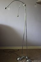Full view of chrome floor lamp approximately 56 inches tall with two articulating eyeball lights at the top attached to curved arms, standing on the floor against a wall.