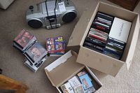 Photo showing a silver and black Sanyo portable stereo with cassette and CD players, a box containing music CDs and DVDs, and some CDs and DVDs spread out next to it on the floor.