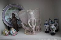 Light-colored large carved elephant statue with three smaller elephants at base, seated Buddha figurines, dark brown standing figurine, and decorative plate.