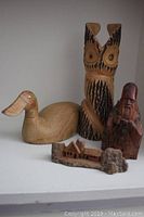 Photo showing wooden duck, stylized owl, man in hat figurine, and miniature carved houses on a white shelf.