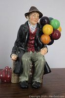 Side view of Royal Doulton Balloon Man figurine seated on crate with cluster of colorful balloons and polka dot bag on floor.