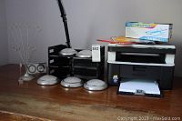 Overview showing Brother DCP 1612W printer with spare toner cartridge box on top, magnifying clamp on light, paper trays, photo clips, push lights, and desk clock on wooden surface.