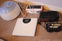 Five devices laid out: white round dome humidifier, orange and white basic digital clock radio, black digital clock radio with red LED, white Taylor lithium digital scale, and white smart clock radio