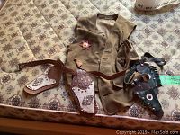 Brown suede cowboy vest laid flat showing front with plastic sheriff badge pinned. Two leather holsters attached to brown belts lying alongside vest on patterned fabric surface.