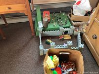 Wooden toy castle with knights on horses displayed on a green base. Adjacent open boxes with toys including cars and trains with visible wear. Carpeted floor background.