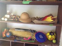Shelf displaying multiple outdoor items including croquet mallets, beige sun hat, yellow and blue balls, blue frisbee, skipping rope, and lawn darts.