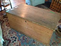 Large wooden dovetailed trunk on patterned rug, showing top and one side with visible wear and natural wood finish.