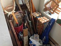 Photo showing collection of vintage sports equipment including wooden croquet mallets, tennis racquets, lacrosse stick, golf clubs, and baseball bats in container and leaning against wall.