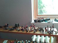 Wide view of assorted toy soldiers and horses displayed on a wooden board near a window, showing variety of figures including British guards and a black cannon.