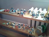 Photo of two shelves filled with approximately 38 Smurf figurines and a small red toy vehicle, displayed on a wooden shelf against a gray wall.