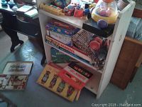 Photo showing wooden three-shelf bookshelf with various board games, puzzles, toys and books stacked and placed on top and around it