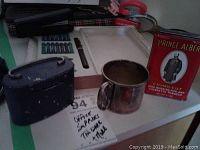 Photo showing silver plated baby cup, Prince Albert tobacco tin, calligraphy nib set with tray, blue leather case, pen holder, and other small office supplies on a white surface.
