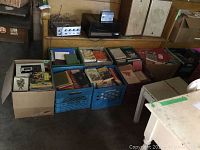 Two cardboard boxes and six milk crates filled with various used books in a dimly lit room near furniture