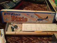 Photo showing Keil Kraft Pixie model airplane kit box, with wood sticks and parts inside, plus detailed plans.