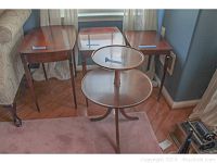 Four wooden tables, including two-tier round pie table in front and three rectangular nesting tables behind, placed in room corner on wood floor.