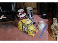 Wide view showing the yellow elephant plant stand, Dresden urn, German Rumtopf pot, Mexican monkey and lizard figure, and Asian ceramic cat.