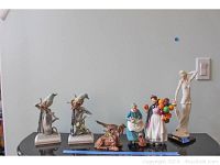 All seven figurines displayed on black surface against wall; includes G. Armani woman with umbrella, Royal Doulton Biddy Pennyfarthing, Andrea Sadek birds, ceramic toad and bighorn.