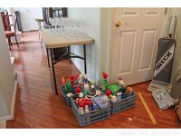 Three bins with assorted cleaning bottles and supplies placed on the floor near a door, with a beige folding table in the background and an Oreck vacuum cleaner visible