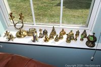 Photo showing 18 solid brass bells of various sizes and designs arranged on a window sill, along with a brass iron box and candleholders at the right end.