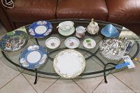 Glass coffee table with assorted decorative plates, bowls, teapot, cups, and collector spoons arranged on top.