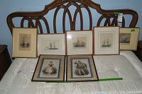 Seven framed art prints displayed on bed with wooden carved headboard; five nautical ship prints and two prints of costumed figures.