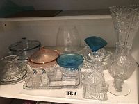 Shelf display of assorted vintage glassware including clear glass covered dishes, a pinkish covered dish, a blue fan-shaped vase, salt and pepper shakers, and clear glass vases and glasses.