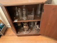 View of 11 glass vases and laboratory style glassware displayed inside a wooden cabinet with two doors open.
