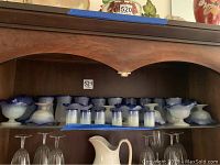 Wooden shelf displaying glassware: 10 frosted glass dessert bowls with a ruffled blue rim and pedestal base, 6 small frosted demitasse glasses with a textured finish, and 10 taller frosted ice tea glasses with blue rims.