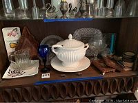 Shelf with white ceramic tureen with lid and ladle, blue glass goblet, clear round serving tray, carved wooden trivet, rectangular and square serving trays, glass coasters.