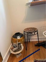 Wide shot showing the plant stand, ceramic vase with repair, and marble stand with rocks on hardwood floor near wall