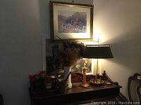 Photo showing the vintage brass lamp turned on, with the hunting scene print above it, brass mortar and pestle, wooden vase, white vase with dried flowers, and candlesticks on a wooden table
