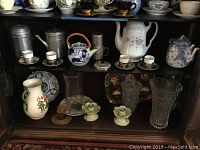 Photo showing multiple items on shelf: aluminum tea set, demitasse cups with saucers, hand painted plates, Swedish vase, crystal vases.