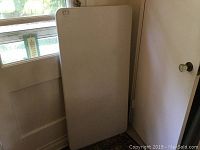 Folded white plastic tabletop leaned against a door, showing the flat smooth top surface with some speckled pattern.
