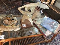 Abstract plaster sculpture placed on glass coffee table with wooden base, seashells on plate, and books visible.