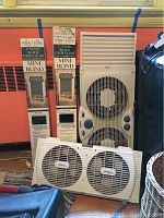 Photo showing two Bradlees vinyl black mini blinds in original packaging, one sealed and one opened, alongside two window fans (Holmes and WindDance) on the floor near a radiator.