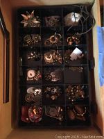 Top view of black tray with multiple small compartments filled with assorted costume jewelry pieces including earrings, pins, and other small accessories.