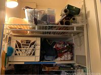 Top shelf with plastic bins holding miscellaneous items including light bulbs, rolls of wrapping paper in boxes, white extension cord hanging