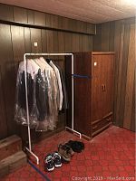 Photo showing white aluminum garment rack with clothes and wooden wardrobe beside it.