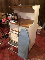 Photo showing white microwave stand with wood-toned shelving, step stool hooked on side, and blue ironing board leaning against it, with clothes steamer on shelf.