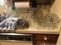Countertop showing various glass pitchers, a martini shaker, juice and rocks glasses, and trays of assorted flatware including spoons and forks.