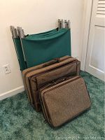 Folded green fabric and aluminum cots standing upright behind two brown tweed suitcases on carpeted floor