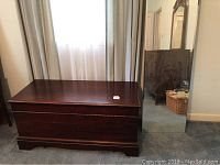 Full view of dark wood Roos cedar chest with push button lid lock, set against a window with curtains, and tall wall mirror to the right.