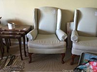 Pair of beige wing back chairs with wooden legs and armrests, placed side by side on carpeted floor, accompanied by small wooden nested tables to the left.