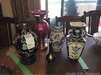 Five vases and candleholder on table including cobalt blue cloisonne vase on wood base, pink glass candleholder, blue Bursleyware pitcher with dragon motif, Noritake floral vase, and small brass vase.