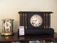 Full lot photo of two clocks on a shelf: one brass anniversary clock with glass dome and one large black mantel clock with gold accents.