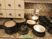 Overall lot showing stacks of plates, bowls, covered casserole, butter dish and mugs on granite countertop