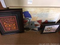 Two framed artworks and one hand-painted canvas artwork leaning against a wall on carpet. Left frame features indigenous style art, center back is unframed abstract painting, right frame shows boats on beach.