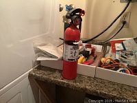 Photo showing a small red fire extinguisher standing on a counter with boxes of miscellaneous tools and hardware parts in background