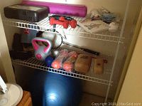 Shelf with exercise equipment including dumbbells, kettlebell, purple yoga mat, wooden weights, badminton racquet, umbrella, and stability ball on the floor.
