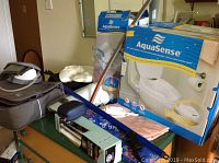 Photo showing multiple health aids and devices on a dresser, including raised toilet seat box, safety rails box, digital blood pressure monitor with cuff, and other items.